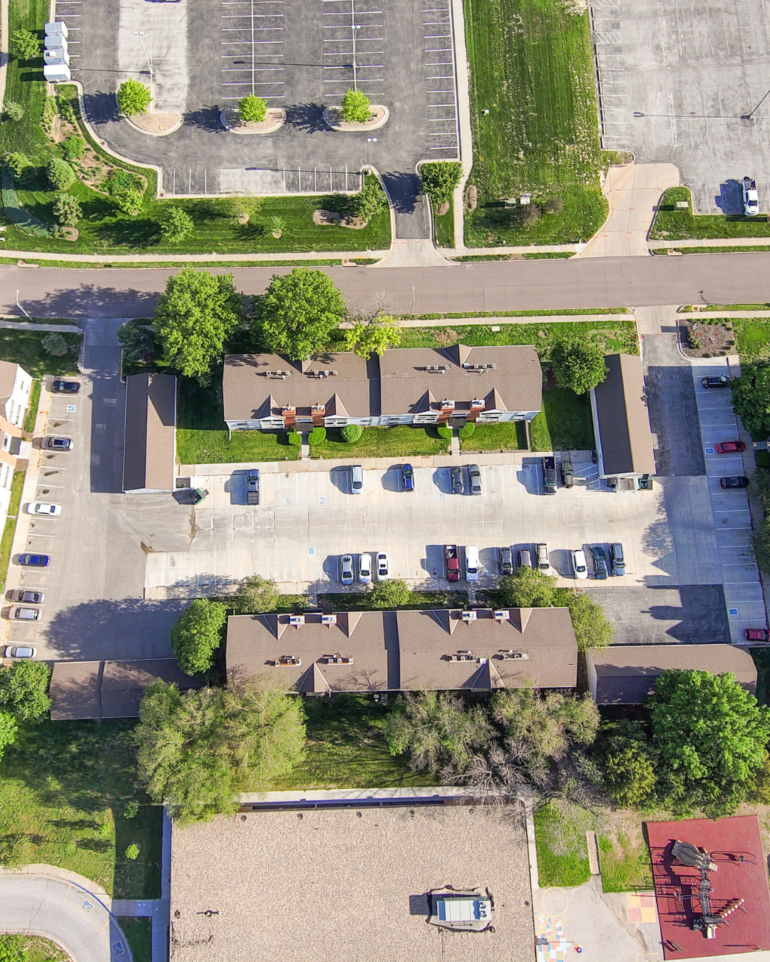 An aerial view of a residential complex with multiple buildings, green trees and a parking lot. Cars are parked along driveways and in the lot. A side-walk and landscaped grassy areas frame the property. The image is captured in daylight with clear visibility of roofs, paths, and surrounding lawns.